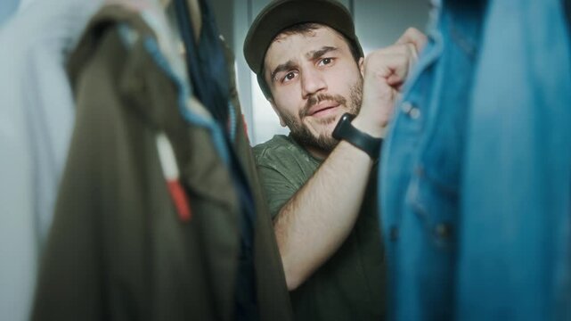 Young man looks indecisive while picking an outfit from closet, unsure what to wear. Concept of decision-making or daily routine.
