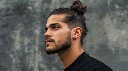A man with a high bun hairstyle, wearing a black t-shirt, standing against a gray concrete wall with a textured surface.