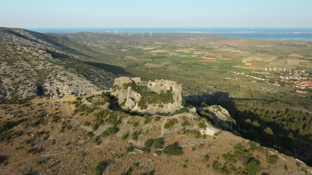 Aerial view of the historic Chateau de Opoul Perillos ruins atop a rugged hill, surrounded by scenic countryside and distant wind turbines in southern France.