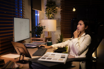 Thoughtful architect sits at her desk reviewing digital blueprints on a monitor while working late in a cozy home office