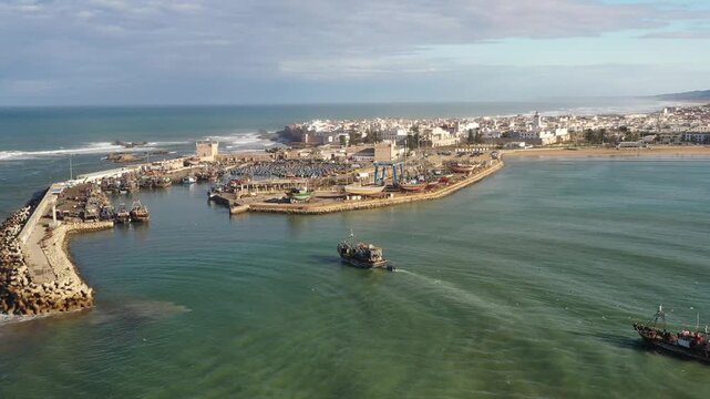 Aerial drone shot flying over the entrance of a busy traditional fishing harbor, showing blue wooden fishing boats docked and navigating the calm water protected by the breakwater