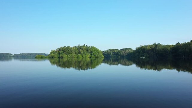 Tranquil lake scene with lush green trees reflecting on the still water under a clear blue sky at Lac des Settons, France.
