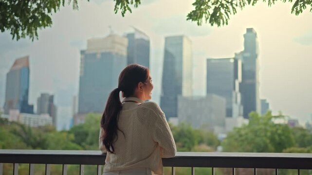 Rear view of a young Asian woman looking at a scenic city skyline with skyscrapers and green park for contemplative urban lifestyle and environmental concept