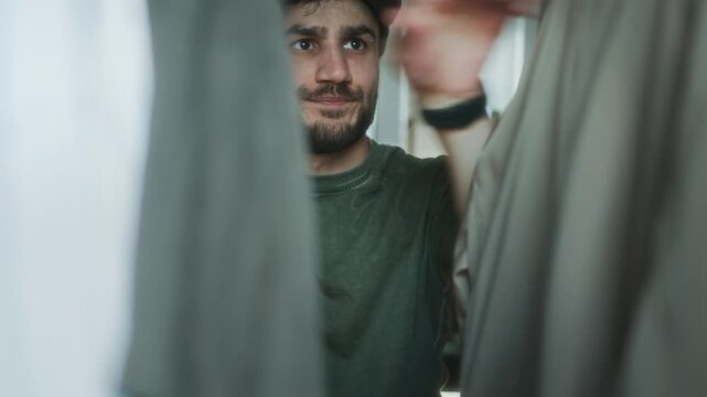 Young man holding a blue shirt against his chest, deciding what to wear as he stands between hanging clothes inside a wardrobe.  
