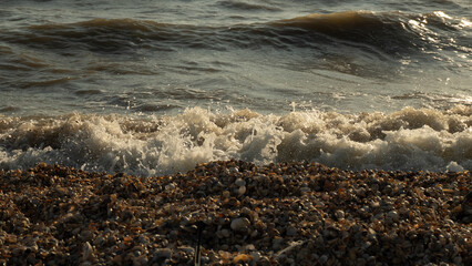 The ocean water turns into white foam and spray. Rolling over the dark pebbles and small shells on the coastal beach, the sea wave creates a dynamic seascape. The sea wave crashes onto the shore.