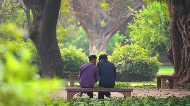 Young indian men sitting together on a park bench leaning on each other enjoying a cozy peaceful morning showing the various forms love and romance can take in the middle of trees and park