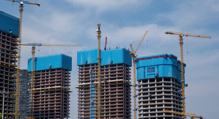 a construction site featuring tall, unfinished skyscrapers with blue protective netting on their rooftops and yellow cranes tower. The buildings have exposed concrete floors and red safety railings