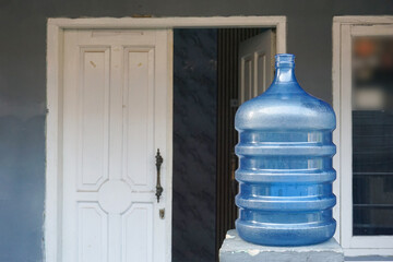 A blue plastic water gallon sits outdoors on a concrete stand, next to a white front door and gray walls. The gallon has a capacity of around 19 liters and is usually used as a household refillable