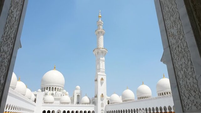 A breathtaking shot frames a grand, pristine white architectural marvel under a clear blue sky. Majestic domes and a towering minaret dominate the scene, exquisitely crafted with intricate details and