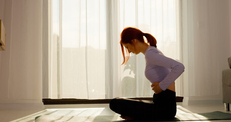 In a living room, a redhead woman sits on a mat, exercising gently while feeling a stomachache. She shows signs of discomfort as she manages her condition.