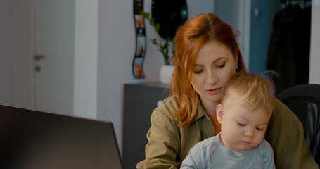 Multitasking redhead businesswoman working remotely, typing on laptop while tenderly holding baby son, balancing career and motherhood in home office setting