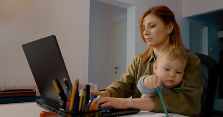 Dedicated working mother multitasking with toddler son on lap, navigating professional responsibilities while providing maternal care and support at home office desk
