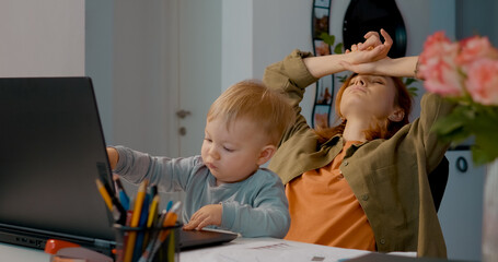 Young child plays with a laptop while the mother takes a break in her home office, surrounded by a workspace filled with colorful stationery and technology.