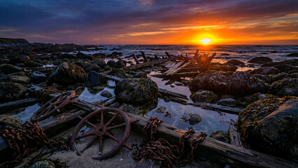Dramatic sunset illuminates the rugged, rocky shoreline scattered with weathered shipwreck debris including rusty ship wheels and heavy iron chains washed ashore.