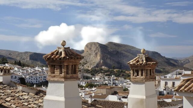 An enchanting panoramic view captures the essence of a traditional hillside village under a serene blue sky dotted with soft clouds. Ornate, traditional brick and terracotta chimneys and tiled rooftop