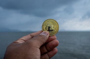 Hand Holding Cryptocurrency Coin Against Ocean and Cloudy Sky, Investment Concept