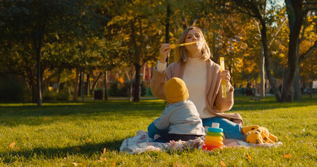 A mother and her young child sit together on a blanket in the park, happily blowing soap bubbles under the warm sun, creating a joyful atmosphere.