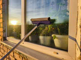 Squeegee wiping a dirty residential window revealing a clear view of a golden sunset and potted plants, symbolizing home cleaning, maintenance, and household chores.