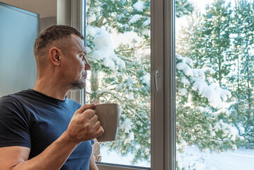 Thoughtful man with mug of hot drink looking out of window to snow scene