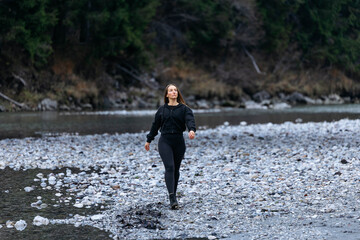 Athletic woman in black activewear walks confidently along a rocky riverbank, surrounded by nature, showcasing a sense of adventure and connection to the outdoors
