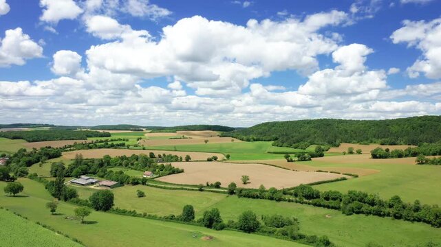 Aerial view of the French countryside with patchwork fields, green trees, and scattered farm buildings under a bright blue sky with fluffy clouds.