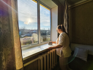 A woman is busy with household chores, wiping a windowsill with a rag. Warm sunlight illuminates the rural landscape outside the window, reflecting the daily routine. The woman is washing the window.