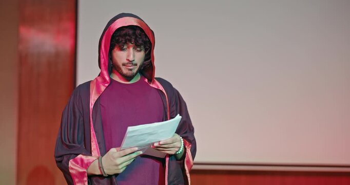 Indian young adult gen z male man standing wear outfit dress hold script paper do play scenes practice indoor hall stage. Teen age boy guy read story line talk speak day time college campus room place