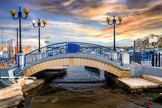 Blue pedestrian bridge in Birzebbuga harbour, Malta, with yellow streetlights, sea view, fishing boats, and traditional houses.