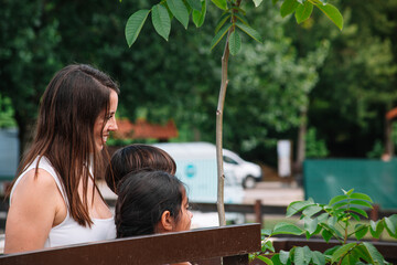 A woman and two children are sitting on a bench in a park. The woman is smiling and looking at the children. The children are looking at something in the distance. The park is surrounded by trees