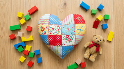 Playful patchwork decorative heart pillow surrounded by colorful wooden blocks and a teddy bear on a light wooden surface
