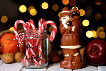 Christmas treats for the holidays, chocolate Santa Claus next to candy canes and healthy fruits with nuts in front of a brightly lit Christmas tree bokeh