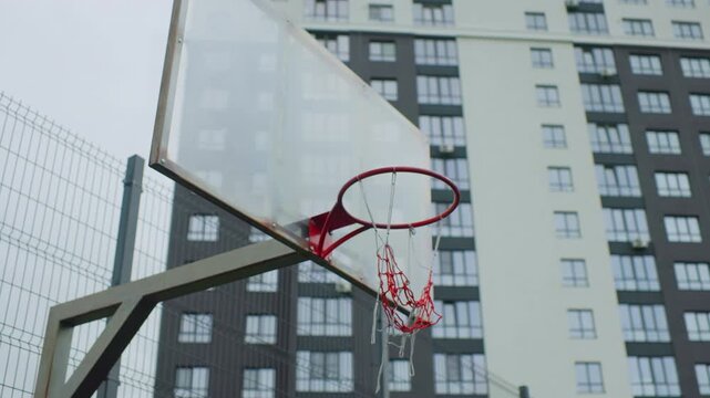 empty basketball hoop against highrise apartments, tattered red net and metal backboard framed by chain mesh fence, overcast sky and concrete pavement evoke quiet solitude and gritty neighborhood