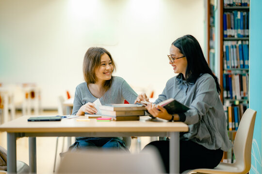 Two female students studying together at library table using tablet and books - Powered by Adobe