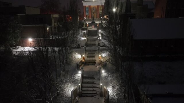 Night drone view of Lynchburg Museum, softly lit by lanterns as fresh snow covers grand stairway and surrounding trees. Calm winter atmosphere in an American historic setting. Approaching rising.