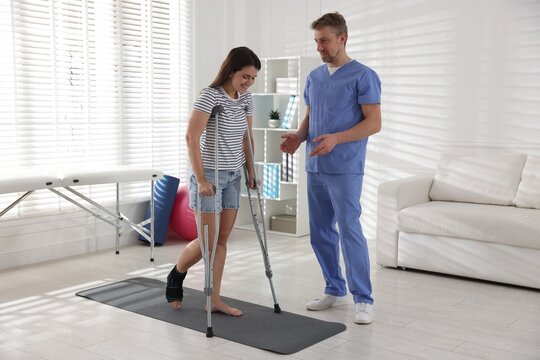 Physiotherapist helping woman with injured leg and axillary crutches during therapy in clinic