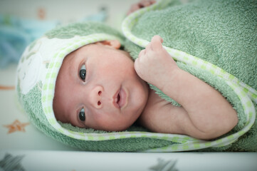 A newborn baby lays on a soft surface, wrapped in a green towel. The baby looks at the camera with curious eyes. The background shows soft colors and designs.