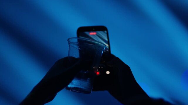 Silhouette hands holding plastic drink cup and smartphone while recording live concert creating nightlife party atmosphere