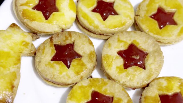 Close-up of homemade Alsatian bredele sandwich cookies with star-shaped red jam filling, golden egg-wash glaze on white plate, festive Christmas holiday dessert