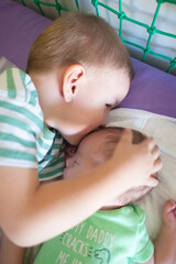 A young boy lies close to his baby sibling on a bed, showing affection by kissing the baby's forehead. They are in a bright room with soft bedding.