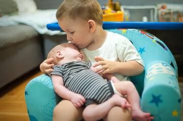 A young child sits on a chair and gently kisses a baby who is resting in their arms. The scene takes place indoors in a home with toys and furniture visible in the background.