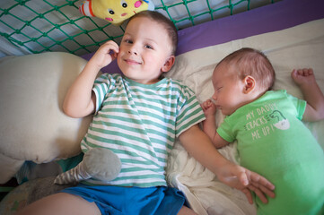 A young boy with short hair lies on a bed with a smile. Next to him, an infant brother sleeps peacefully, surrounded by soft toys. Sunlight comes through the window.