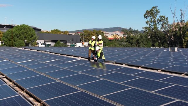 Three engineers wearing safety vests and hard hats collaborating on a solar panel maintenance check on an industrial rooftop on a sunny day - Powered by Adobe