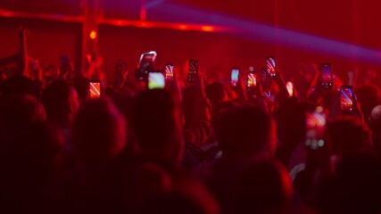 Slow motion audience jumping and dancing while filming live concert as stage lights flicker and create energetic immersive atmosphere