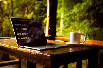 Laptop and cup on wooden table of terrace with tropical forest view at sunset, remote work digital nomad