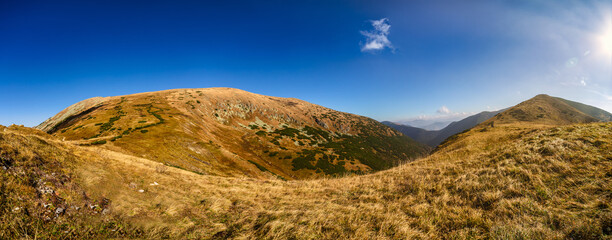 A wide panoramic view of a rocky hiking trail winding through the slopes of the Low Tatras National Park during a sunny autumn day. Beautiful yellow grass and blue sky
