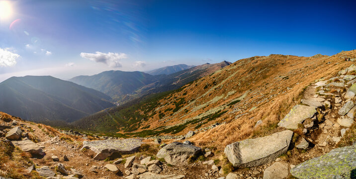 A wide panoramic view of a rocky hiking trail winding through the slopes of the Low Tatras National Park during a sunny autumn day. Beautiful yellow grass and blue sky