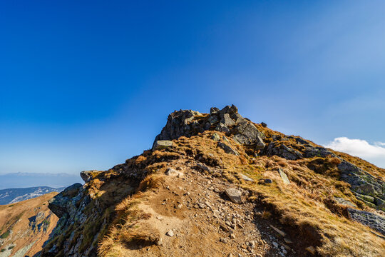 A wide panoramic view of a rocky hiking trail winding through the slopes of the Low Tatras National Park during a sunny autumn day. Beautiful yellow grass and blue sky