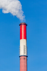 A tall, red and white industrial chimney stands against a vibrant blue sky, releasing a plume of smoke.