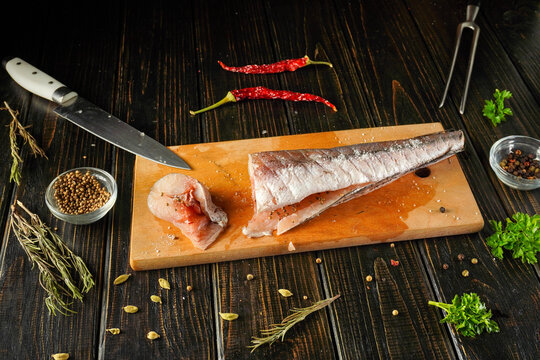 A Merluccius chef cuts a fish on a wooden board. There are spices and herbs scattered around. A knife rests nearby while red chili peppers add color to the scene