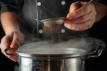 A chef holds a fine mesh strainer over a pot in a kitchen. Steam rises as the chef carefully adds spices or paprika to enhance the flavor of the dish